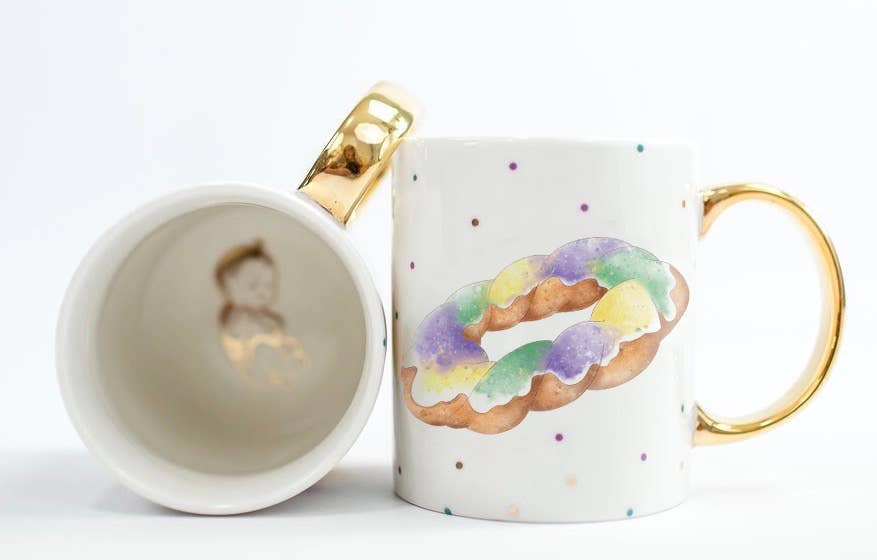 White mug with gold handle and painted king cake beside a second mug showing a gold baby inside