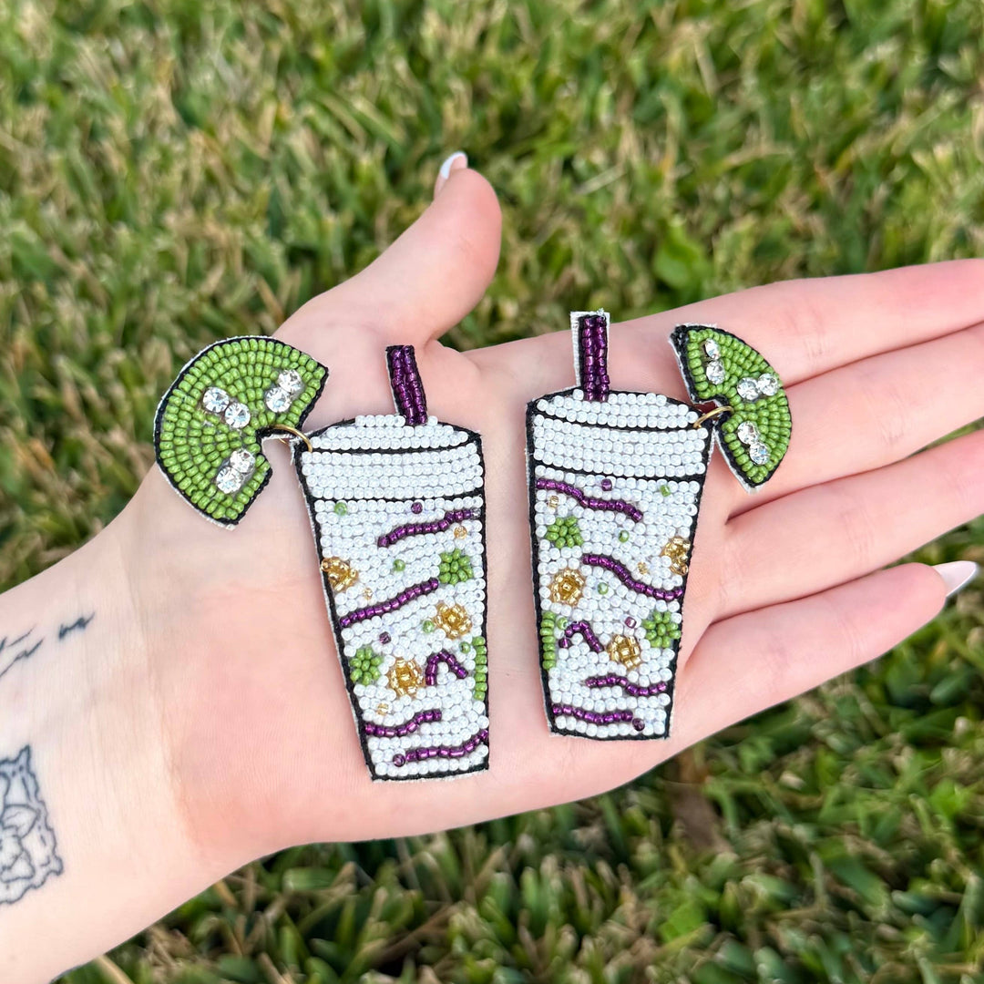 Hand-beaded cocktail earrings held in a palm over green grass featuring vibrant purple straws, lime wedges with sparkling rhinestones, and festive green and gold bead patterns