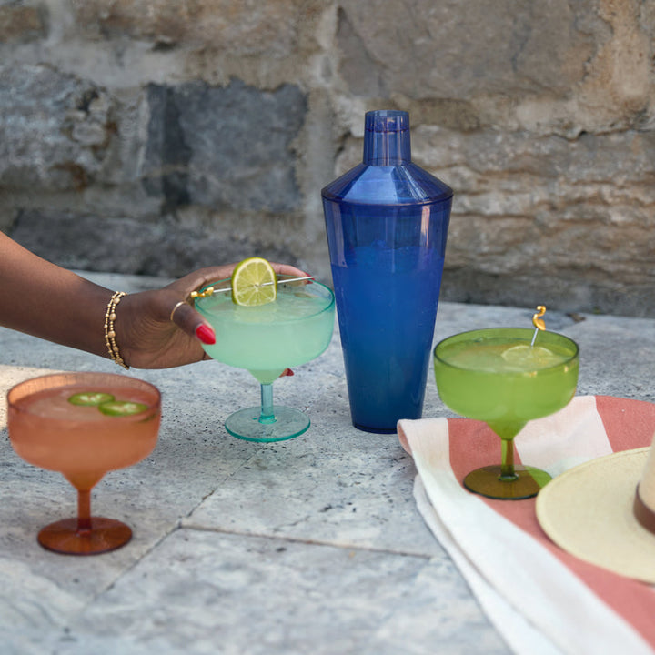 Photograph of vibrant summer cocktails and a blue translucent shaker on a stone patio featuring a hand with red nails holding a lime-garnished margarita, a pink drink with sliced jalapeños, and a green cocktail accented by a golden flamingo stirrer
