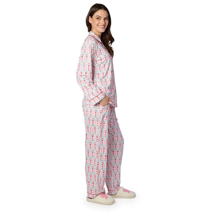 Digital studio photograph of a woman modeling a nutcracker-themed pajama set against a white background, highlighting the red-piped trim, light blue wreath pattern with pink bows, and cozy cream slippers with matching pink ribbon details