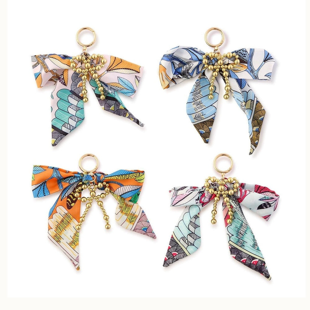 Fabric and gold-tone metal accessory charms featuring four vibrantly patterned silk ribbon bows in orange, teal, and pink botanical prints adorned with central gold-beaded hearts and dangling bead chains on a white background