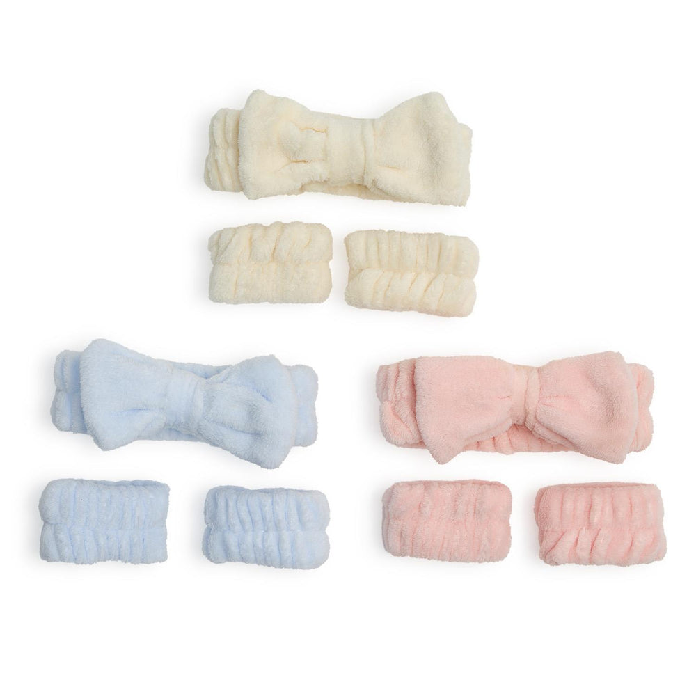 Studio photograph of three spa sets consisting of plush bow headbands and matching wristbands in cream, light blue, and soft pink colors arranged against a white background featuring a fuzzy microfiber texture and large decorative bows