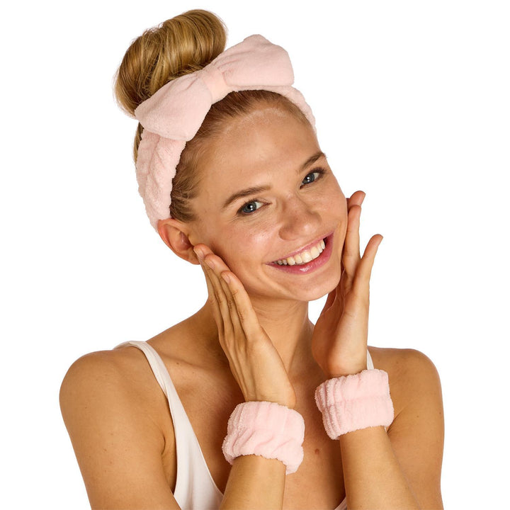 Studio photograph of a smiling blonde woman with a high bun wearing a light pink plush bow headband and matching fuzzy wristbands against a plain white background
