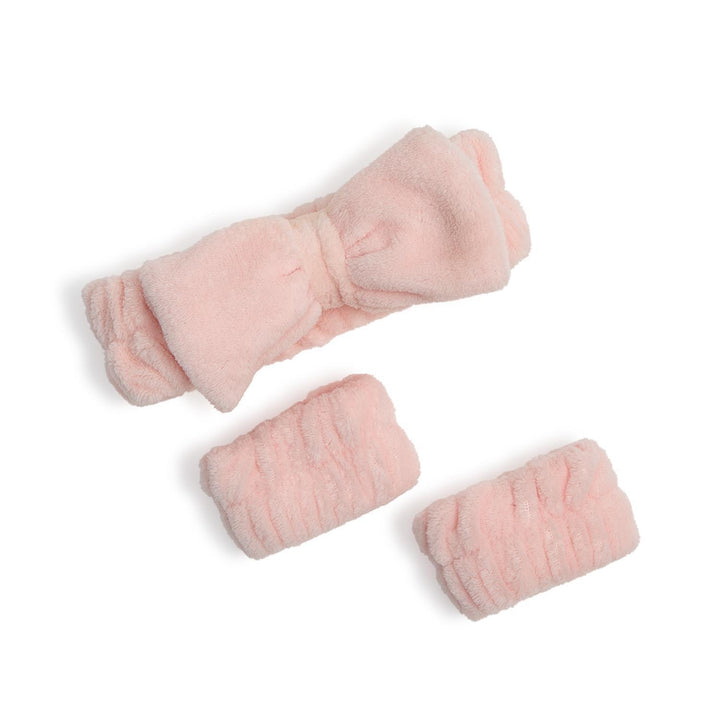Studio product photography of a light pink three-piece skincare set featuring a plush microfiber headband with an oversized bow and two matching ruched wristbands on a white background