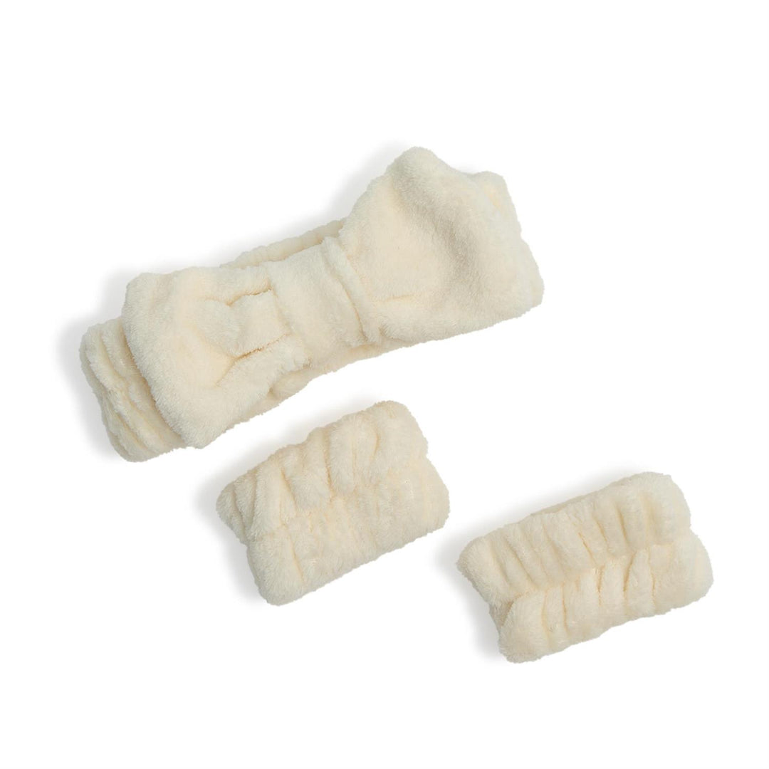 Studio product photograph of an ivory microfiber spa set consisting of a headband with a large central bow and two matching wristbands on a plain white background featuring a soft plush texture and gathered elasticized design