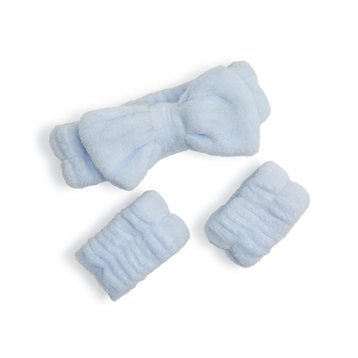 Studio product photograph of a light blue plush skincare set featuring a prominent bow-adorned headband and two matching ribbed wristbands with a soft microfiber texture on a plain white background