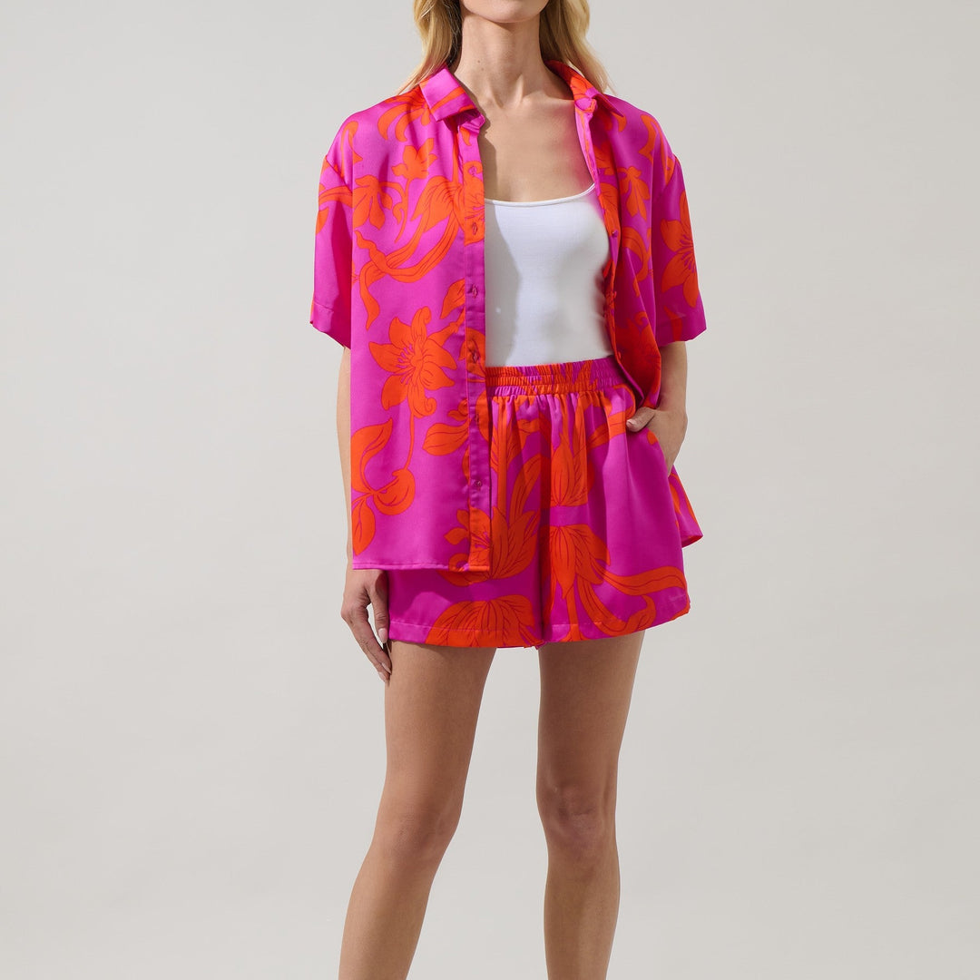 Studio photography of a female model in a minimalist studio wearing a bold magenta and orange tropical floral matching set composed of a short-sleeved button-down shirt and high-waisted shorts over a white top