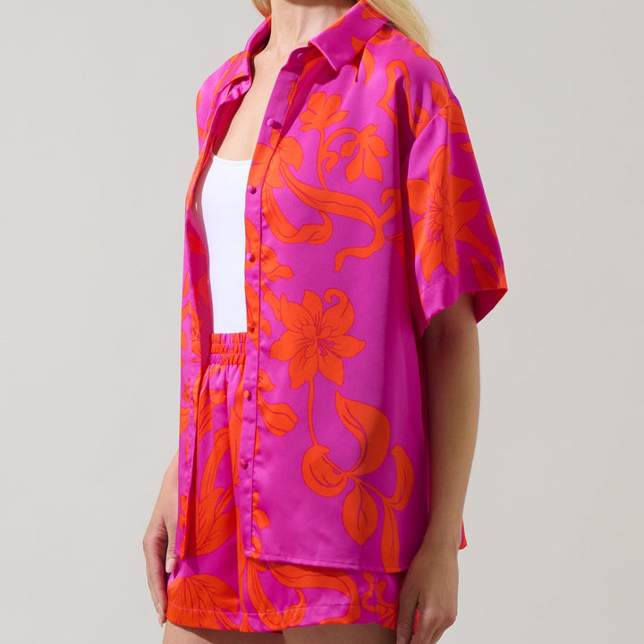 Digital studio photography of a woman wearing a vibrant magenta and orange floral matching set featuring a short-sleeved button-down shirt with large lily patterns on satin fabric against a neutral backdrop