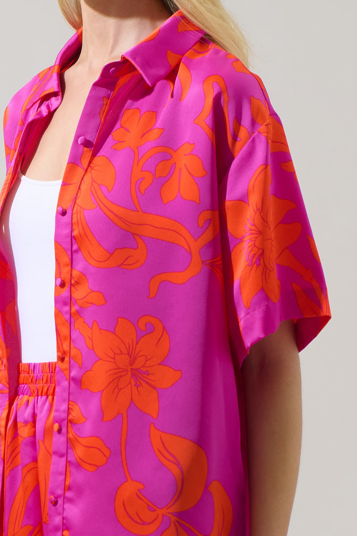 Studio fashion photograph of a woman wearing a vibrant fuchsia pink and orange floral print short-sleeve shirt with matching shorts featuring large lilies and fabric-covered buttons against a plain gray background