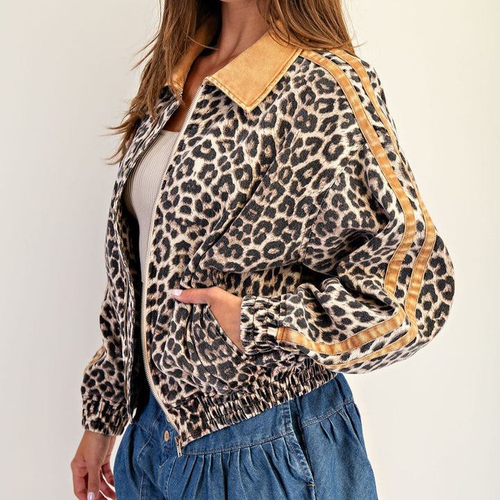 Photograph of a woman modeling a leopard print bomber jacket featuring a tan corduroy collar and triple tan stripes along the sleeves paired with a blue denim skirt against a plain studio background