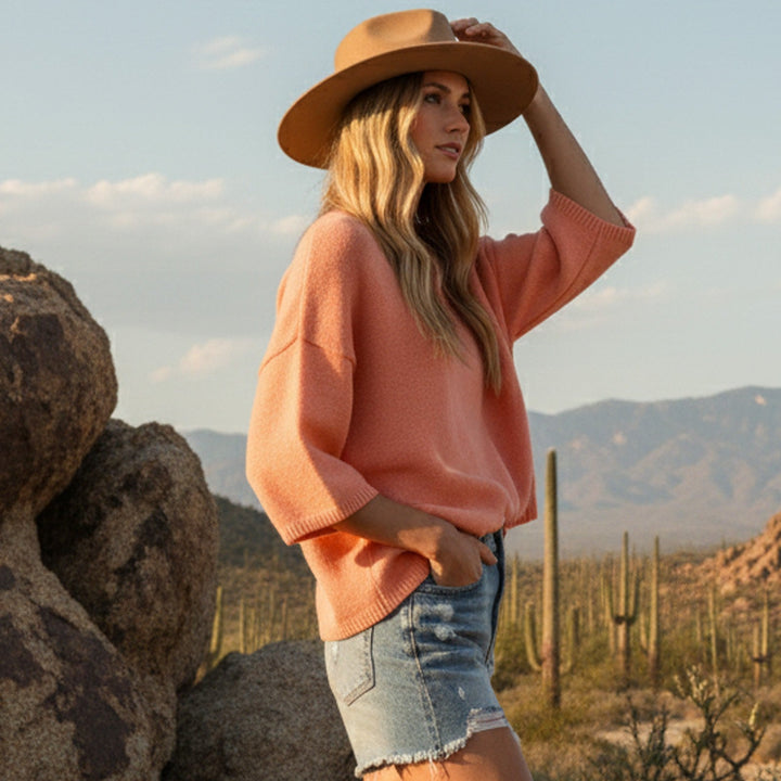 Photograph of a woman wearing a wide-brimmed tan felt hat and a peach-colored knit sweater posing in the Sonoran Desert among iconic saguaro cacti and weathered rock formations