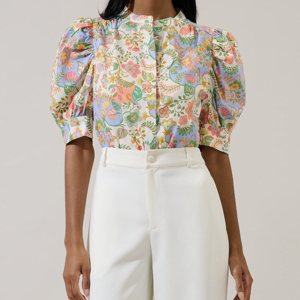 Photograph of a woman in a studio wearing a colorful floral puff-sleeve blouse featuring pink and blue patterns and white high-waisted trousers