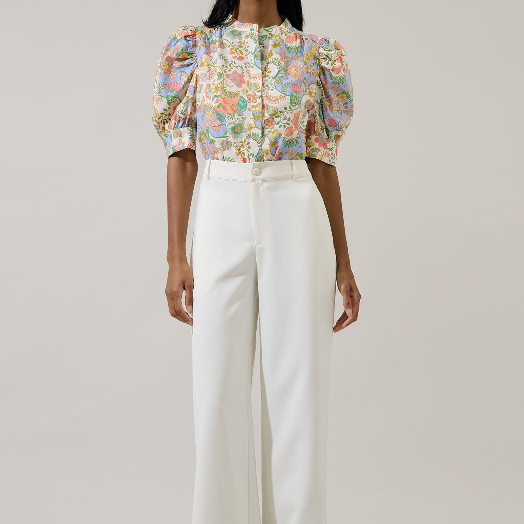 Photograph of a fashion model in a studio wearing a pastel floral-print blouse with puffed sleeves and orange botanical motifs tucked into structured white high-waisted trousers