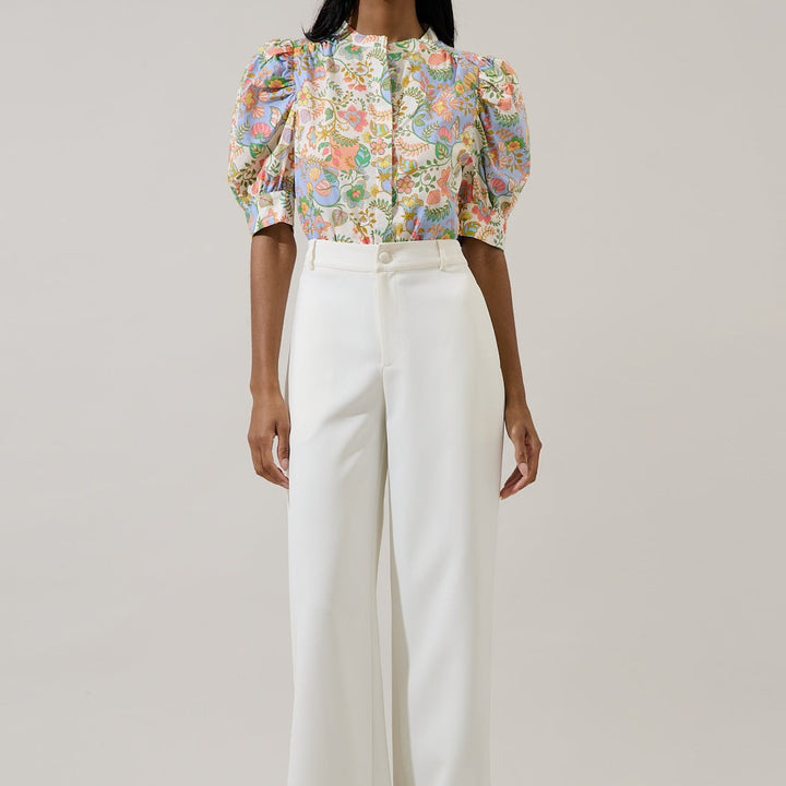Photograph of a fashion model in a studio wearing a pastel floral-print blouse with puffed sleeves and orange botanical motifs tucked into structured white high-waisted trousers
