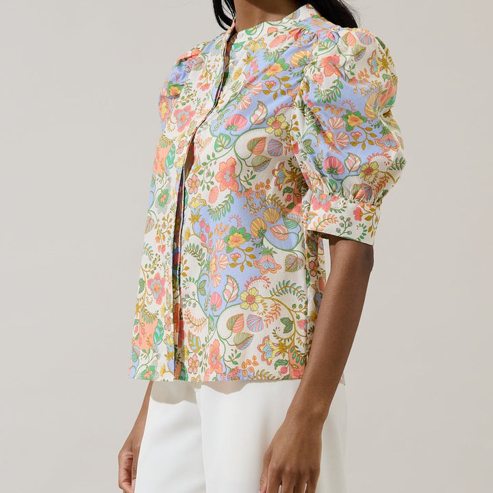 Photograph of a woman wearing a vibrant floral print blouse featuring puffed elbow-length sleeves, a mandarin collar, and intricate botanical patterns in pastel pink, blue, and yellow against a neutral studio background