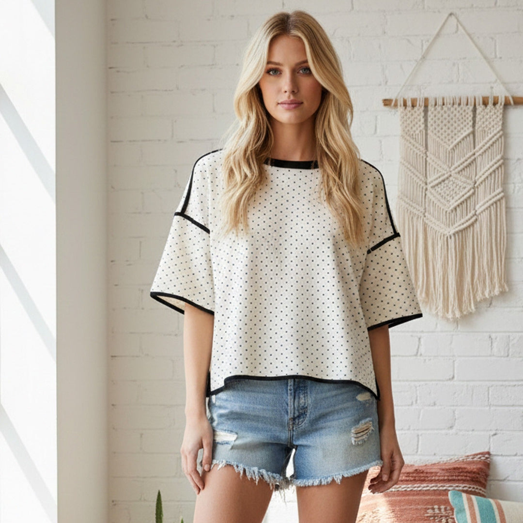 Photograph of a blonde woman in a bright indoor studio with a white brick wall wearing a cream polka dot top with black piping and distressed denim shorts next to a fringed macramé wall hanging