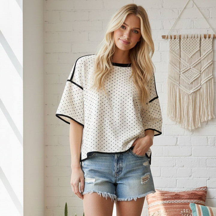 Photograph of a blonde woman in a white brick room featuring a cream macrame wall hanging, wearing a black-trimmed white polka-dot top and distressed denim shorts with frayed hems