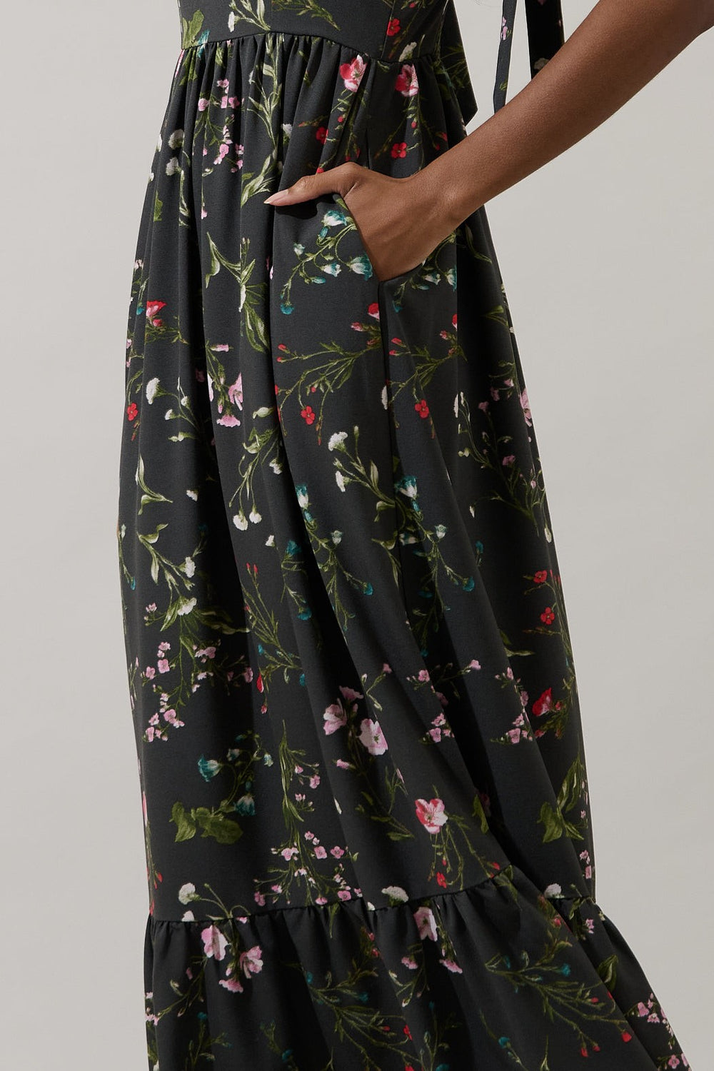 Digital photograph of a person wearing a black maxi dress with delicate pink and white floral prints featuring a functional side pocket and a ruffled bottom hem against a plain grey background