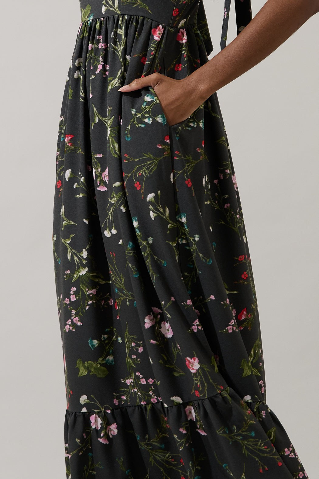 Digital photograph of a person wearing a black maxi dress with delicate pink and white floral prints featuring a functional side pocket and a ruffled bottom hem against a plain grey background