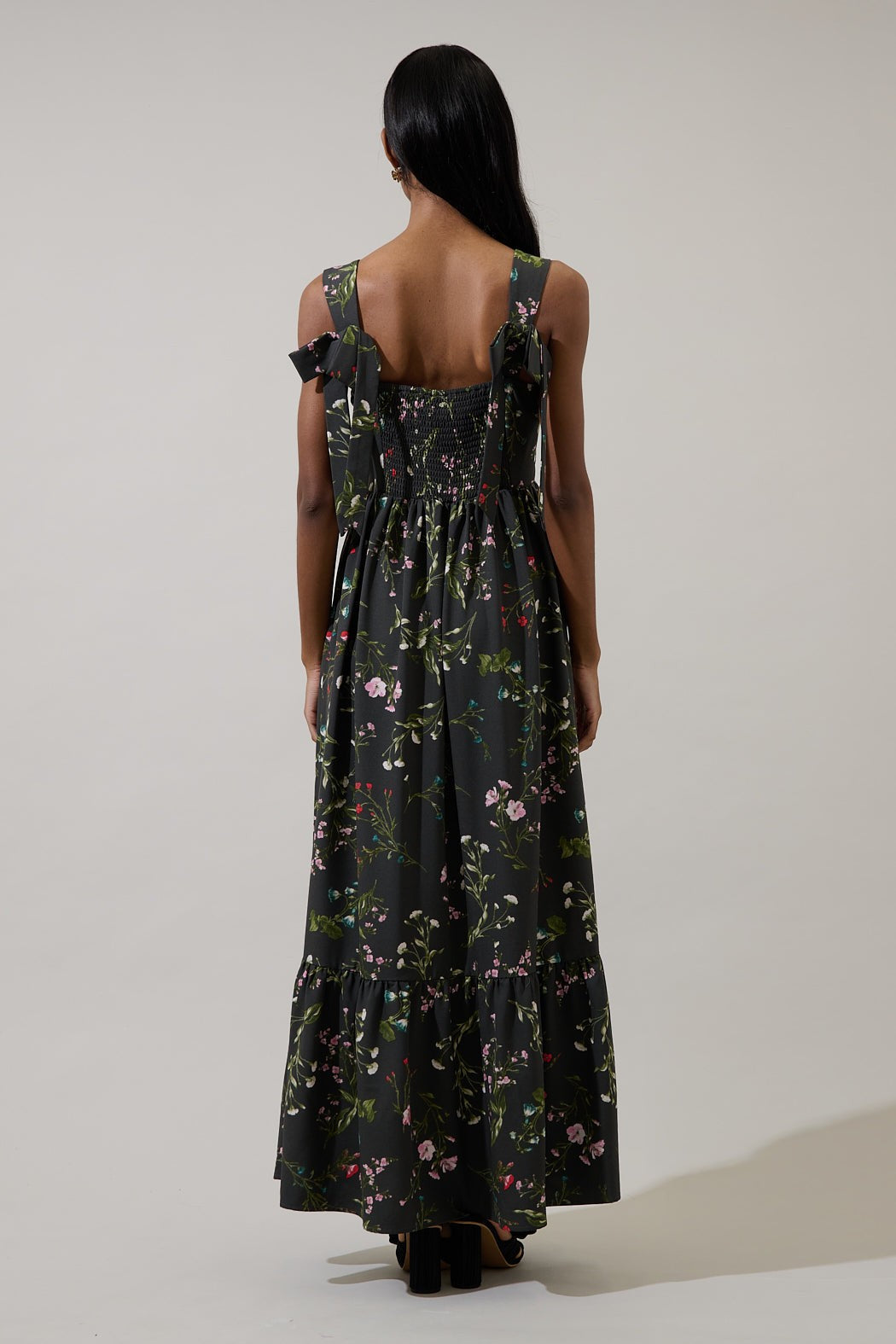 Fashion photograph of a model viewed from behind in a studio wearing a black maxi dress featuring a vibrant floral print, bow-tie shoulder straps, and a smocked back panel