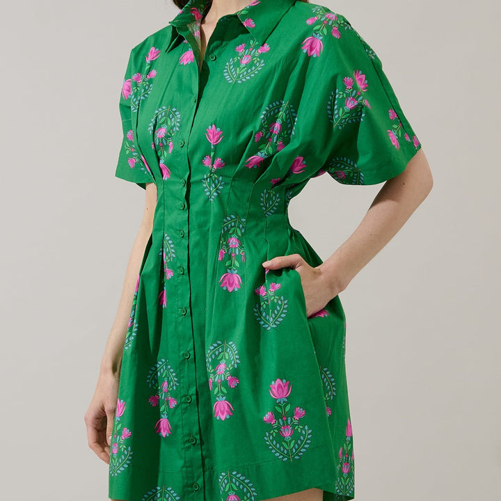 Fashion photograph of a model wearing a vibrant emerald green shirtdress with pink floral block prints in a studio, featuring a cinched pleated waist and functional side pockets
