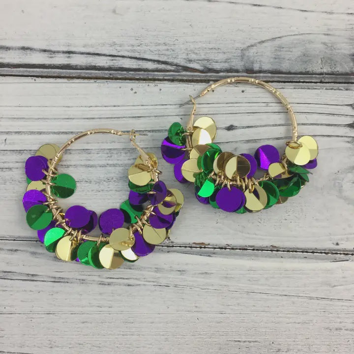 Handmade jewelry of Mardi Gras hoop earrings featuring shimmering purple, green, and gold metallic sequins clustered on gold-toned circular frames resting on a white distressed wooden surface