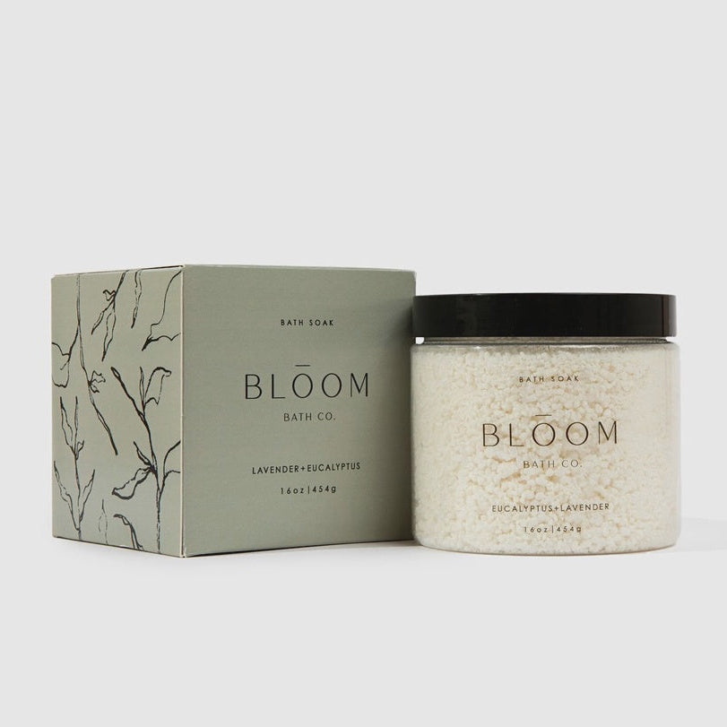 Product photograph of Bloom Bath Co. Lavender and Eucalyptus bath soak in a neutral studio setting featuring a muted sage green box with botanical line drawings and a transparent jar of coarse white mineral salts with a matte black lid