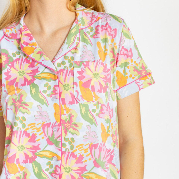 Photograph of a woman wearing a short-sleeved pajama top featuring a vibrant pink, yellow, and green watercolor-style floral print, bright pink piping along the notched lapels, and small white buttons against a neutral white background