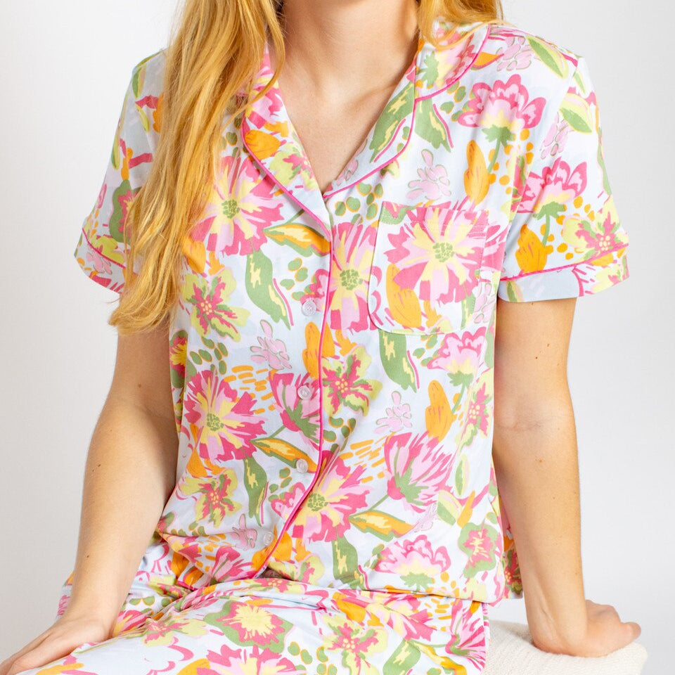 A studio photograph of a woman wearing a short-sleeved button-up pajama top featuring a vibrant pink and yellow floral pattern with hot pink piping and a small chest pocket against a neutral background