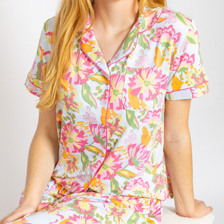A studio photograph of a woman wearing a short-sleeved button-up pajama top featuring a vibrant pink and yellow floral pattern with hot pink piping and a small chest pocket against a neutral background