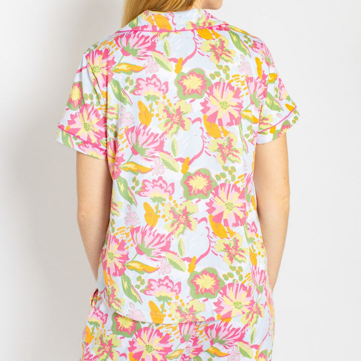 Photograph of a woman from the back wearing a Lilly Pulitzer style floral pajama top featuring vibrant pink and yellow blooms with green accents and bright pink piping against a plain white background
