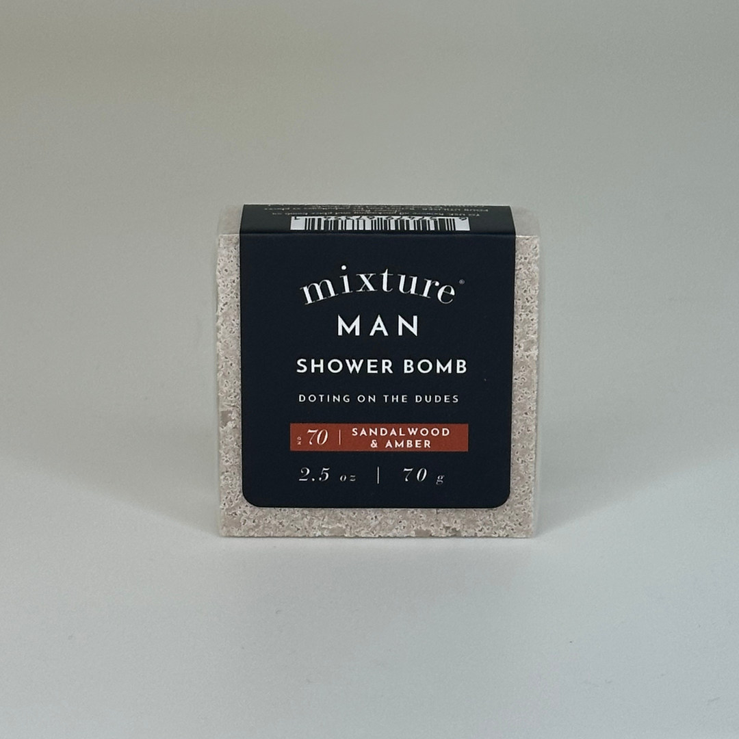 Product photograph by Mixture of a square Mixture Man Sandalwood &amp; Amber shower bomb featuring a speckled granite-like texture, a matte black wrap-around label, and a burnt orange fragrance bar against a neutral gray background
