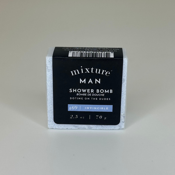 Product photography of a Mixture Man shower bomb in a studio setting, showcasing its square white crystalline texture through clear wrapping and a black label with bold white typography and a light blue scent bar for No. 69 Invincible