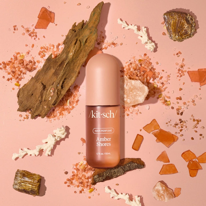 Commercial studio photograph by Kitsch of a frosted Amber Shores hair perfume bottle centered on a soft pink backdrop featuring textured driftwood, white coral branches, and scattered grains of pink sea salt