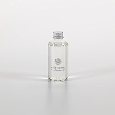 Studio photograph of a clear glass bottle of White Hibiscus Home Fragrance Oil featuring a silver metal screw cap and a delicate floral emblem while casting a soft reflection on a minimalist white surface
