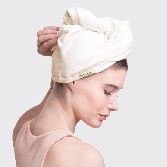 Commercial photograph by Kitsch of a woman in a peach tank top wearing a cream-colored microfiber hair towel wrap featuring a small black woven logo tag against a neutral studio background