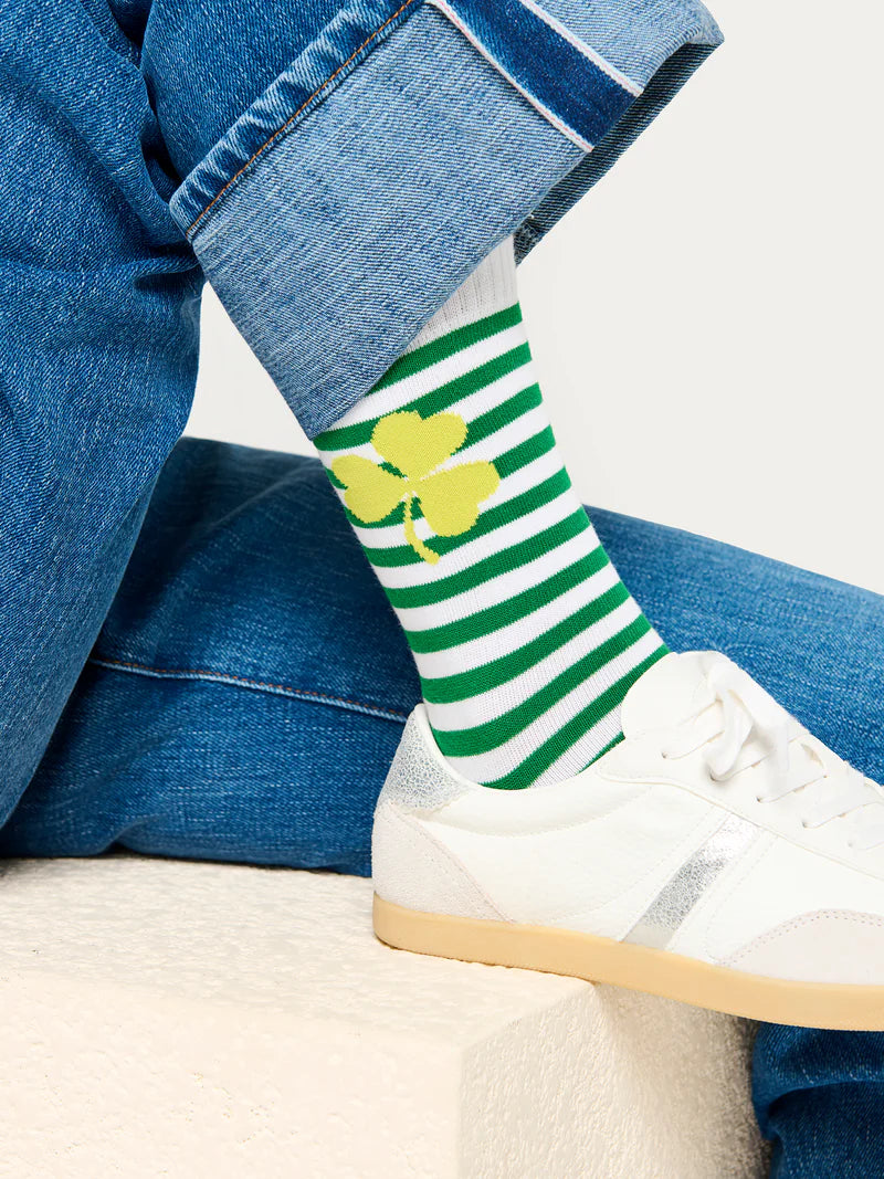 A studio photograph of a person's ankle resting on a white stone ledge showcasing cuffed blue denim jeans and white sneakers paired with green and white horizontal striped socks featuring a vibrant yellow shamrock