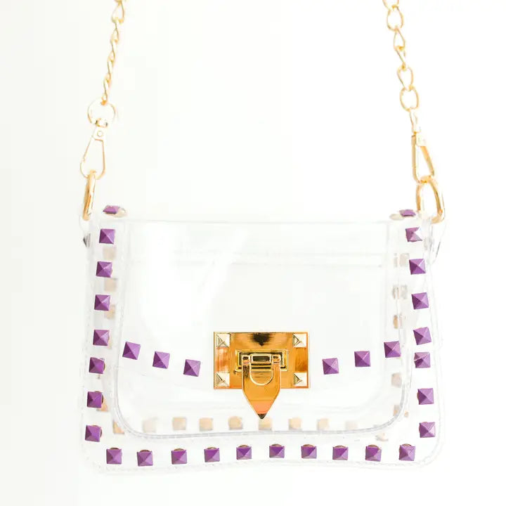 Digital photograph of a transparent PVC crossbody bag featuring vibrant purple pyramid studs, a gold-tone turn-lock clasp, and a gold-link chain strap against a minimalist white background