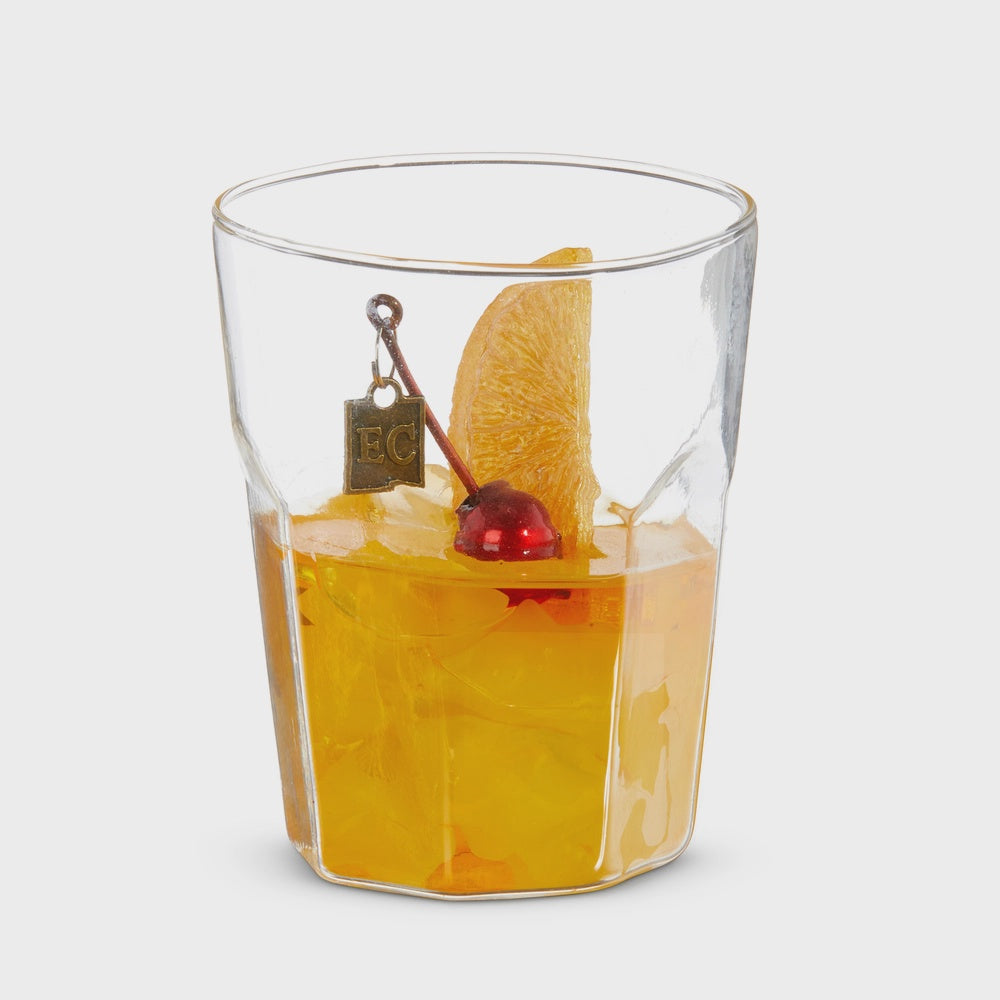 Glass sculpture by Eric Cahan of an Old Fashioned cocktail set in a studio environment featuring a faceted glass, a bright red maraschino cherry, and a textured orange slice adorned with a signature gold-colored EC metal tag