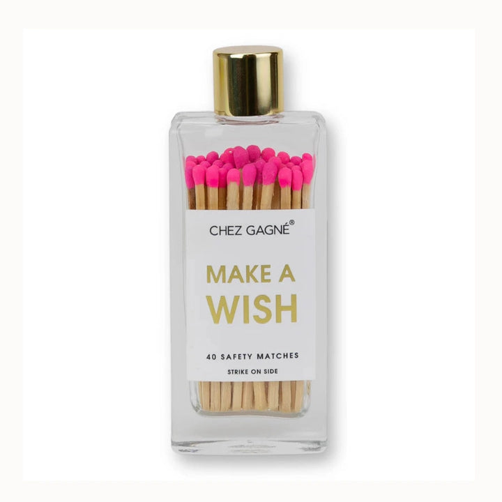 Product photograph by Chez Gagné of a clear glass apothecary bottle containing safety matches with vibrant hot pink tips, featuring a metallic gold cap and gold foil Make A Wish text against a clean white background