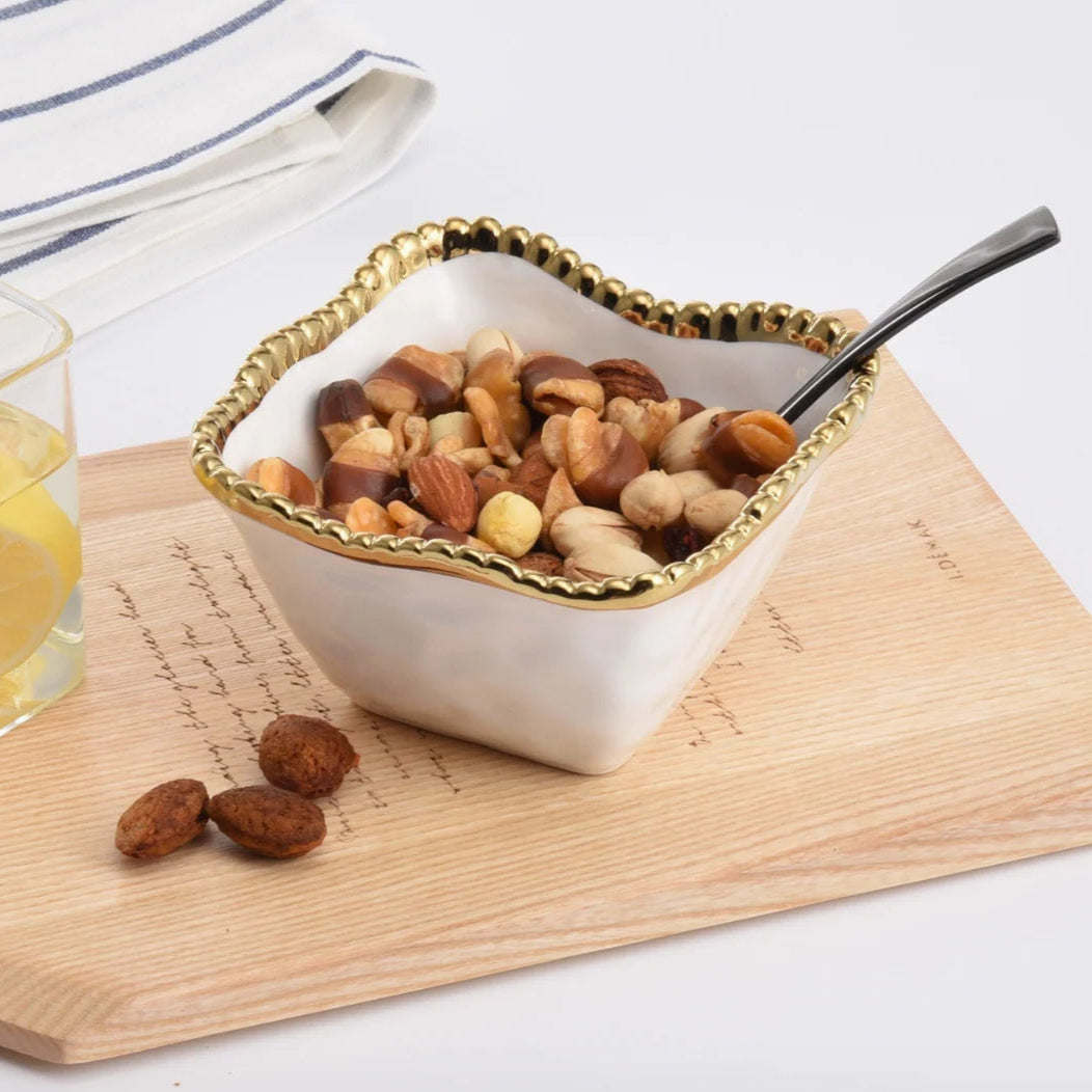 Ceramic white bowl with a scalloped gold rim filled with mixed nuts on a wooden platter featuring handwritten script alongside a black spoon and a glass of lemon water
