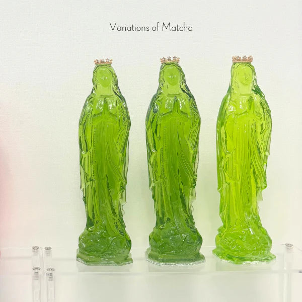 Translucent glass sculptures of the Virgin Mary on a transparent shelf featuring three figures in varying shades of matcha green with tiny gold crowns and hands clasped in prayer