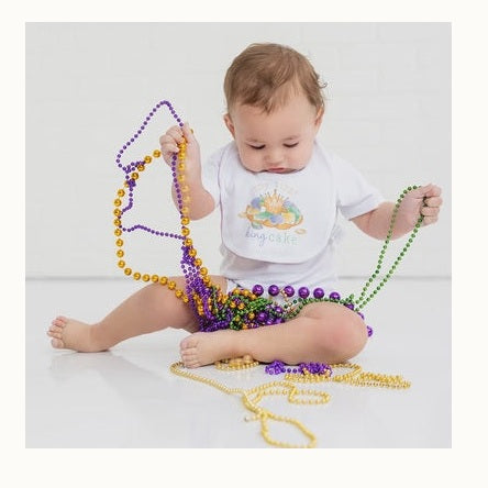 Digital photograph of a baby sitting on a white floor surrounded by vibrant purple, green, and gold Mardi Gras beads while wearing a white bib with a king cake illustration and the text My First King Cake