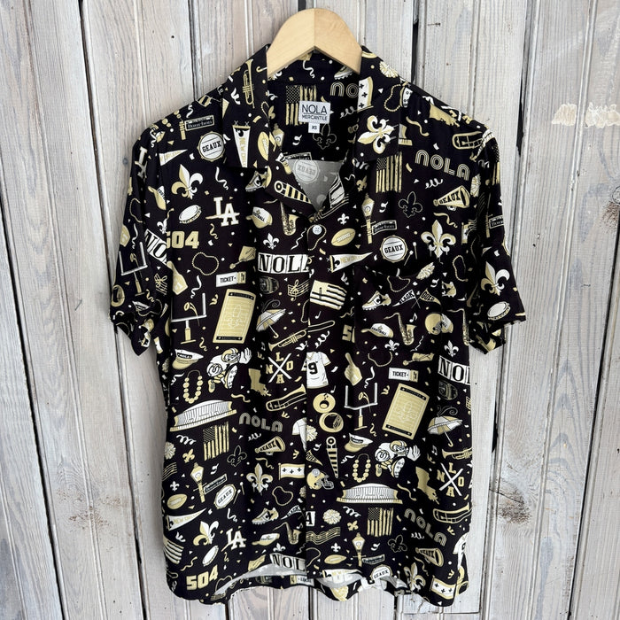 Photograph of a NOLA Mercantile short-sleeve button-down shirt featuring a black and gold New Orleans-themed pattern with the Caesars Superdome, Bourbon Street signs, fleur-de-lis symbols, and jazz instruments hanging against a rustic wooden wall