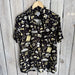 Photograph of a NOLA Mercantile short-sleeve button-down shirt featuring a black and gold New Orleans-themed pattern with the Caesars Superdome, Bourbon Street signs, fleur-de-lis symbols, and jazz instruments hanging against a rustic wooden wall