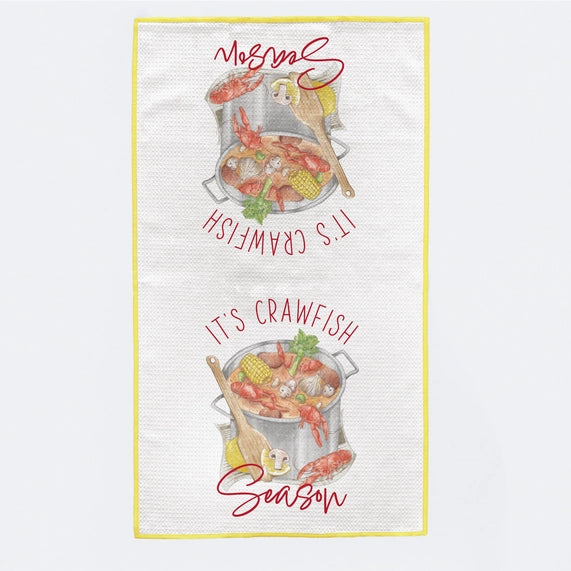 Graphic illustration on a white waffle-weave kitchen towel featuring two mirrored stockpots filled with vibrant red crawfish and yellow corn on the cob framed by the red text IT'S CRAWFISH SEASON and a bright yellow border