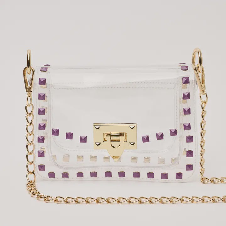 Product photograph of a clear PVC crossbody bag featuring purple pyramid rockstuds, a gold-tone chain strap, and a matching turn-lock closure against a plain studio background