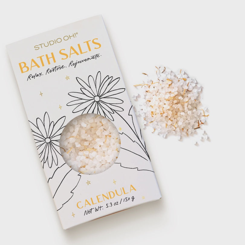 Product photography by Studio Oh! of Calendula bath salts on a white surface featuring a box with black line-art flowers, yellow typography, and coarse white crystals mixed with vibrant orange dried petals