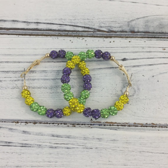 Beaded jewelry featuring a pair of interlocking-style hoop earrings in traditional New Orleans Mardi Gras colors on a white-washed wood background, showcasing vibrant purple, green, and gold textured beads and gold-finished metal components