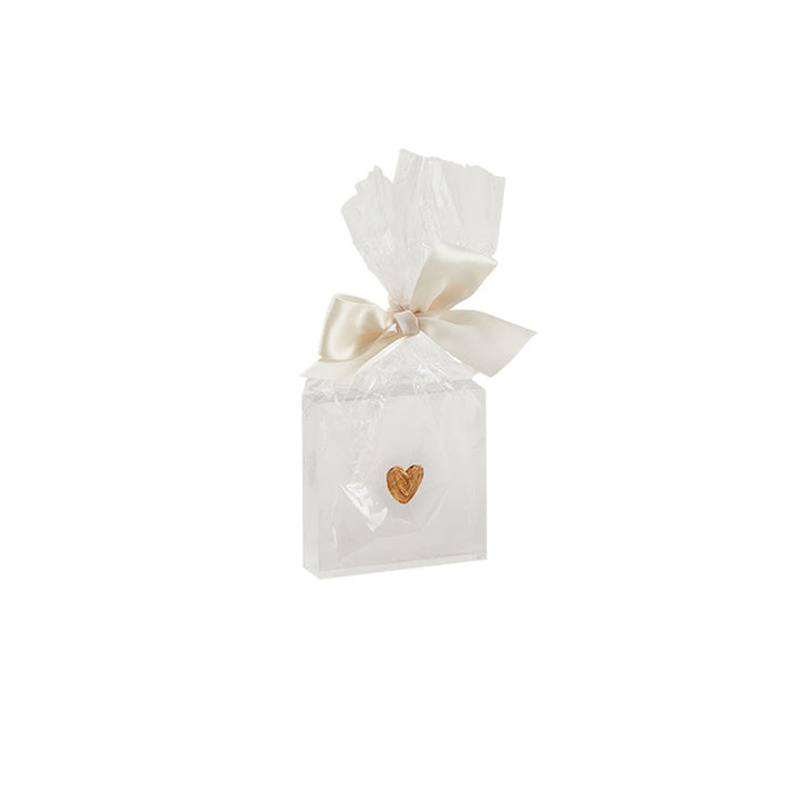 Studio photograph of a square white gift favor wrapped in clear crinkled cellophane with a small textured gold heart charm and tied with an elegant cream satin ribbon bow on a plain white background